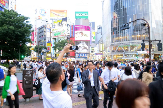 Eine Gruppe von Menschen, die auf einem Weg gehen, wobei ein Mann ein Mobiltelefon hält, umgeben von Bäumen, Pfählen, Verkehrssignalen, Gebäuden und Schildern mit Text.