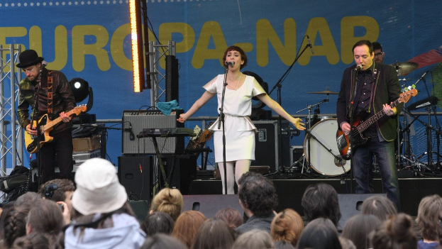 Vier Personen, die auf einer Bühne Musik machen, mit einer Frau in einem weißen Kleid, die singt, und zwei Männern, die Gitarren spielen, während das Publikum darunter eine Wand mit Lautsprechern im Hintergrund sieht.