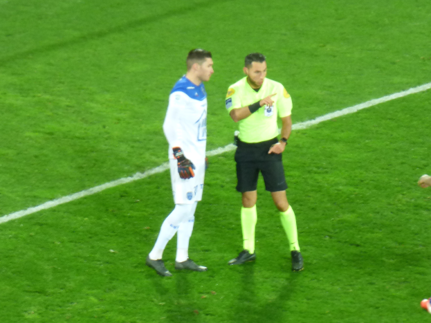Ein Schiedsrichter in gelbem T-Shirt und schwarzen Shorts spricht mit einem Füßballspieler in weißer und blauer Uniform auf dem Feld, beide tragen Turnschuhe.