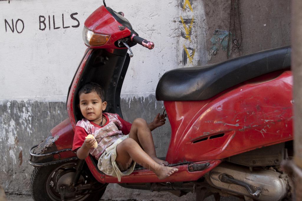 Ein Junge sitzt auf einem Roller, h√§lt ein Objekt in der Hand, vor einer Wand mit Schrift darauf.
