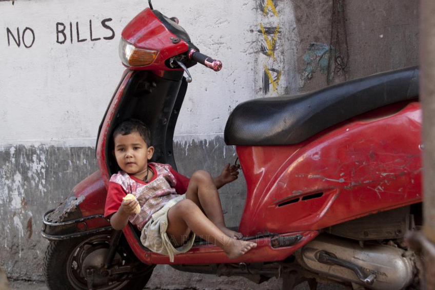 Ein Junge sitzt auf einem Roller, h√§lt ein Objekt in der Hand, vor einer Wand mit Schrift darauf.