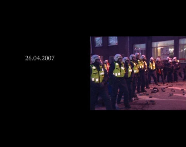 Gruppe von Polizisten in Helmen und Jacken vor einem Gebäude mit Fenstern stehend, mit einem sichtbaren Datum auf der linken Seite.