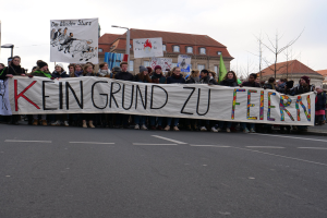 Demonstranten mit einem Transparent "Kein Grund zu Feiern" gegen deutsche Sparmaßnahmen, mit Straßeninfrastruktur und Gebäuden im Hintergrund.