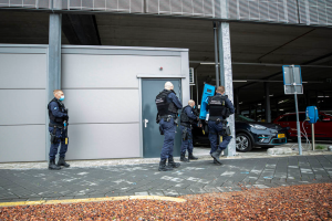 Eine Gruppe von Polizeibeamten steht vor einem Gebäude mit einer Tür, umgeben von Fahrzeugen, Pfählen, Brettern und anderen Gegenständen auf einem Parkplatz mit verstreuten Steinen auf dem Boden.
