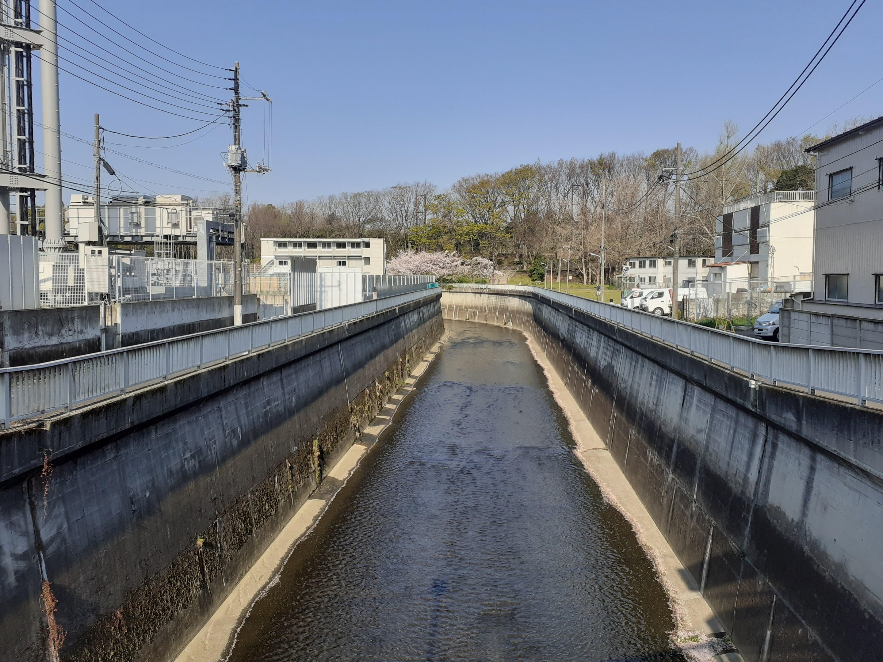 Ein kleiner Kanal verläuft durch eine Stadt mit Gebäuden auf beiden Seiten, Strommasten mit Drähten und Fahrzeugen auf der Straße, mit Bäumen und einem klaren blauen Himmel im Hintergrund.