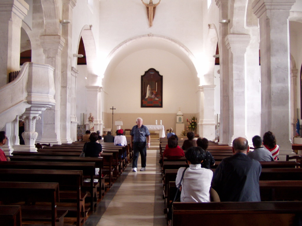 Ein alter Mann geht einen Mittelgang in einer Kirche entlang, flankiert von Sitzreihen mit sitzenden Individuen, während die Wände im Hintergrund Fotografien und Idole zeigen.