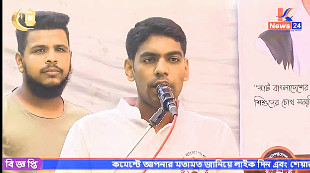 Zwei Männer stehen nebeneinander vor einem Fernseher, einer spricht in ein Mikrofon in einem weißen Hemd, der andere in einem aschfarbenen T-Shirt, mit einem Banner und Logo dahinter und einem Watermark unten.