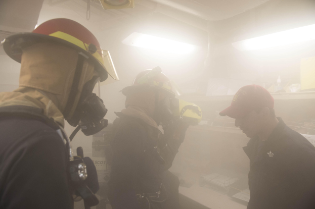 Feuerwehrleute in Helmen mit Rauch aus dem Mund, einer hält eine Kamera, während einer Trainingsrunde mit Beleuchtung und Equipment im Hintergrund.