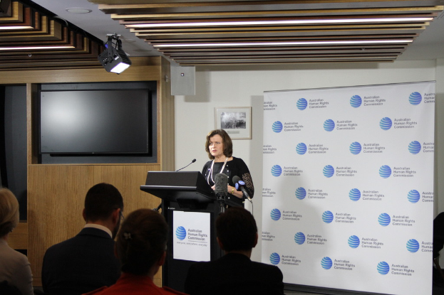 Eine Frau steht auf einer Bühne und spricht in ein Mikrofon, vor ihr sitzen Menschen auf Stühlen, an der Wand hängt ein Banner, ein Fernseher an der Holzwand und Lampen an der Decke.