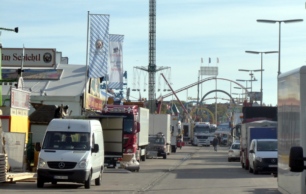 Vollgestellte Straße mit Lastwagen, Autos, Fußgängern, Laternen, Bannern, einem Turm, einer Attraktion, Bäumen und einem bewölkten Himmel.