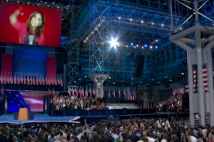 Eine große Menschenmenge sitzt vor einer Bühne mit einem Rednerpult, Mikrofon, Fahnen, einem Bildschirm mit einer Frau und Lichtausrüstung, wahrscheinlich bei einer Präsidentschaftsdebatte.