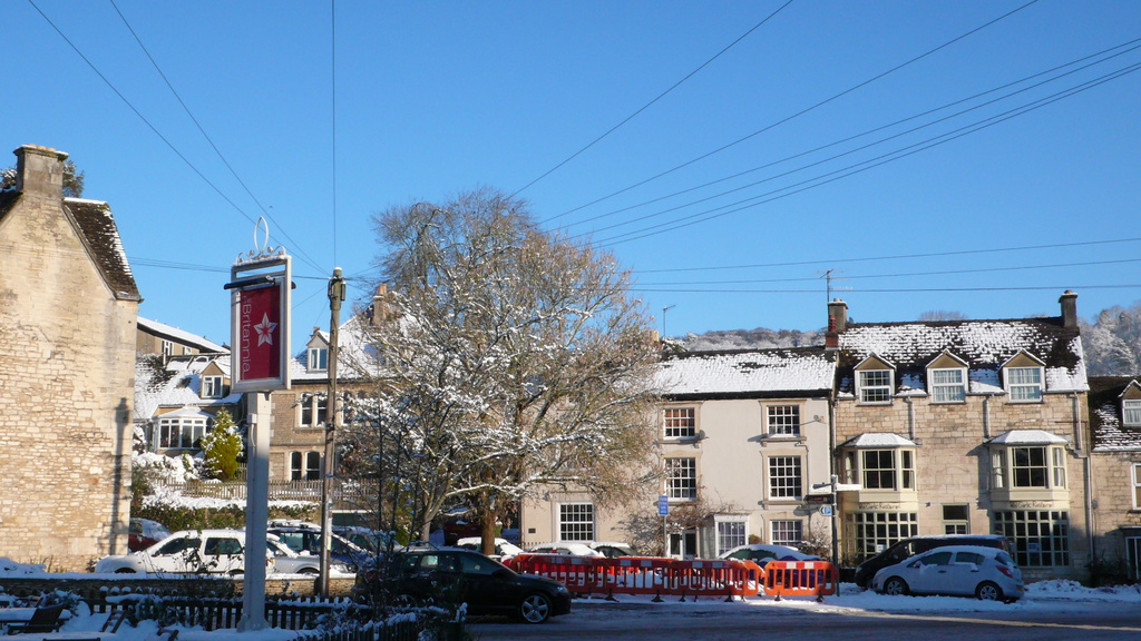 Fahrzeuge auf einer schneebedeckten Straße mit Häusern, Bäumen und Strommasten im Hintergrund.