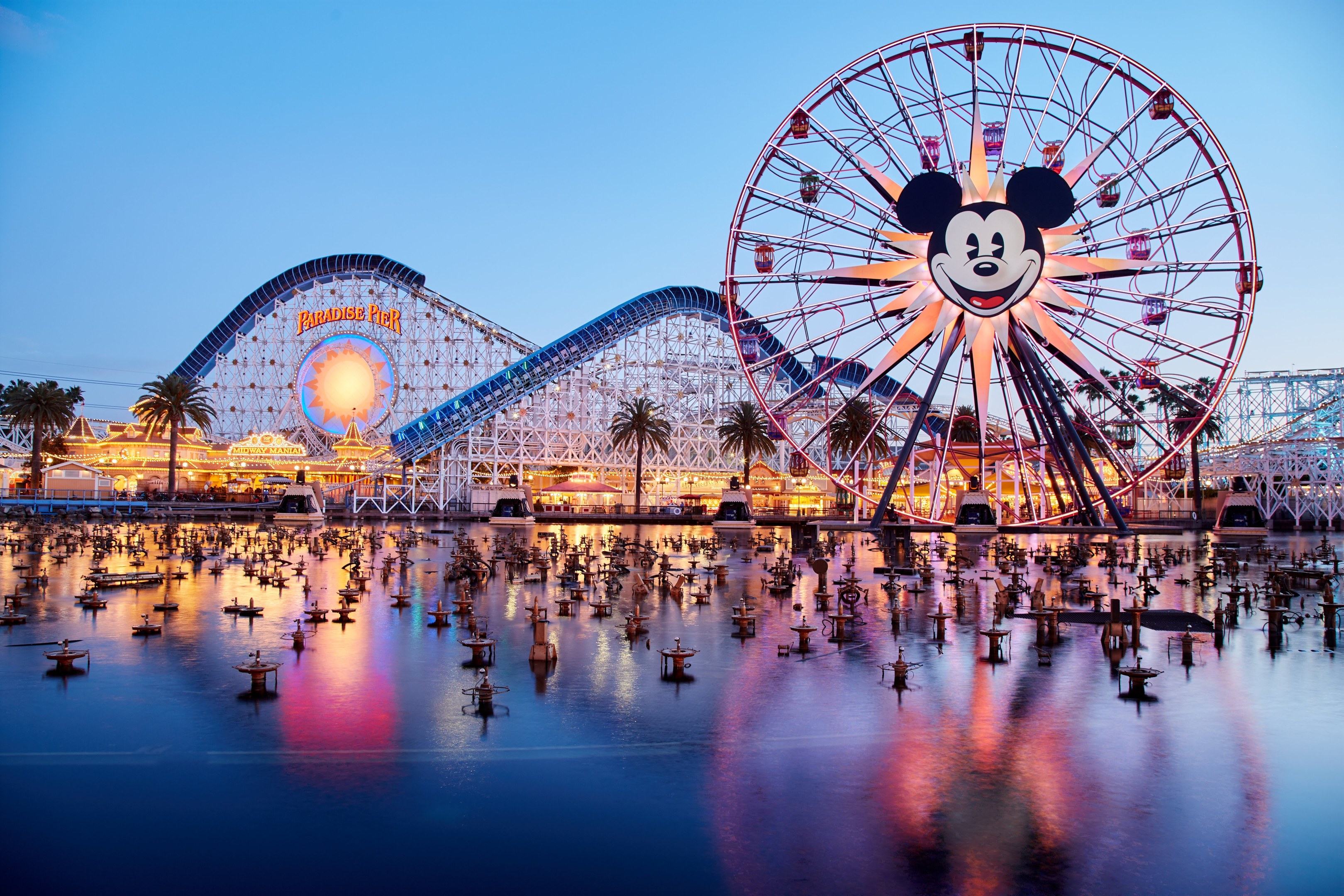 Eine lebendige Dämmerungsszene im Disneyland California Adventure mit einem riesigen Mickey-Maus-Rad im Vordergrund, einer Achterbahn im Hintergrund und Booten auf dem Wasser, mit einem farbenfrohen Himmel und silhouettenhaften Bäumen.