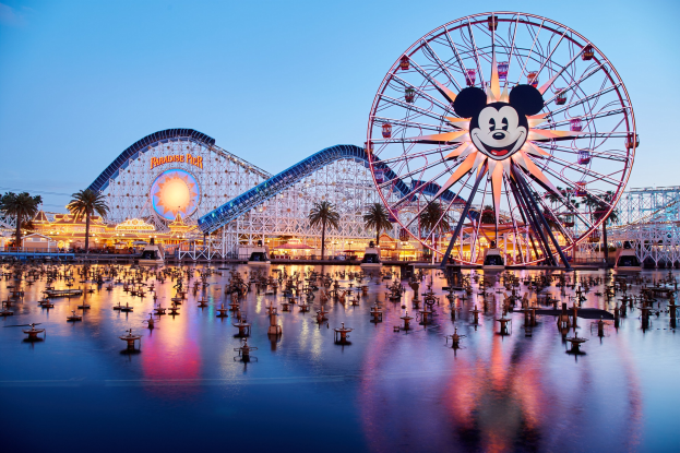 Eine lebendige Dämmerungsszene im Disneyland California Adventure mit einem riesigen Mickey-Maus-Rad im Vordergrund, einer Achterbahn im Hintergrund und Booten auf dem Wasser, mit einem farbenfrohen Himmel und silhouettenhaften Bäumen.