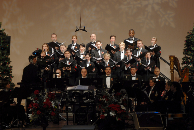 Eine Gruppe von Menschen auf einer Bühne mit Bücher haltend, umgeben von Musikinstrumenten, Blumenbouquets und Weihnachtsbäumen mit Lichtern und einem Bildschirm im Hintergrund.