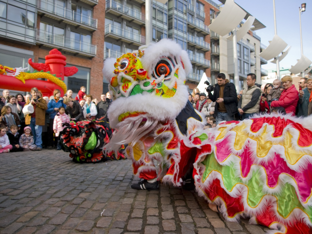Vibrierendes chinesisches Neujahrsfest in Amsterdam mit einer Drachenvorführung und einer Zuschauermenge, einige machen Fotos, vor einer Kulisse aus Gebäuden, Laternenmästen und einem klaren blauen Himmel.
