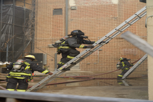 Eine Gruppe von Feuerwehrleuten in Helmen und Zylindern klettert an einer Leiter vor einem Backsteingebäude hoch, mit Rohren und einer Metallstange auf dem Boden und einem Gebäude mit Fenstern und einem Netz im Hintergrund.