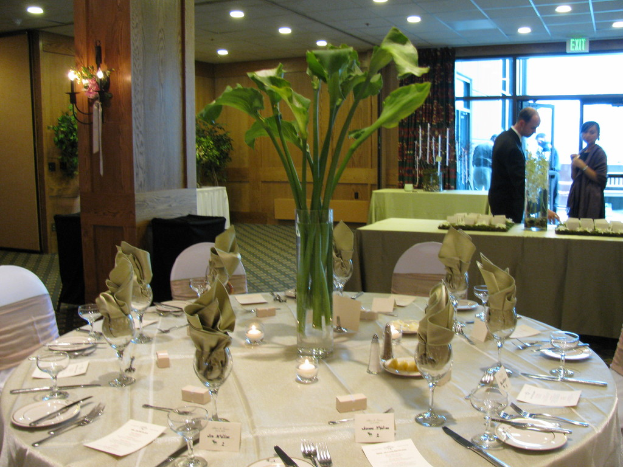 Ein Tisch mit einer weißen Decke, Tellern, Besteck und einem Glas, umgeben von Stühlen, mit einem grünen Tisch dahinter. Ein Mann in einem schwarzen Blazer und ein Mädchen stehen in der Nähe einer Ausgangstür, mit zwei Männern, die draußen stehen.