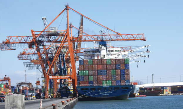 Eine Hafenszene mit großen Kränen, Waren auf einer Brücke neben dem Meer und Schiffen im Hintergrund.