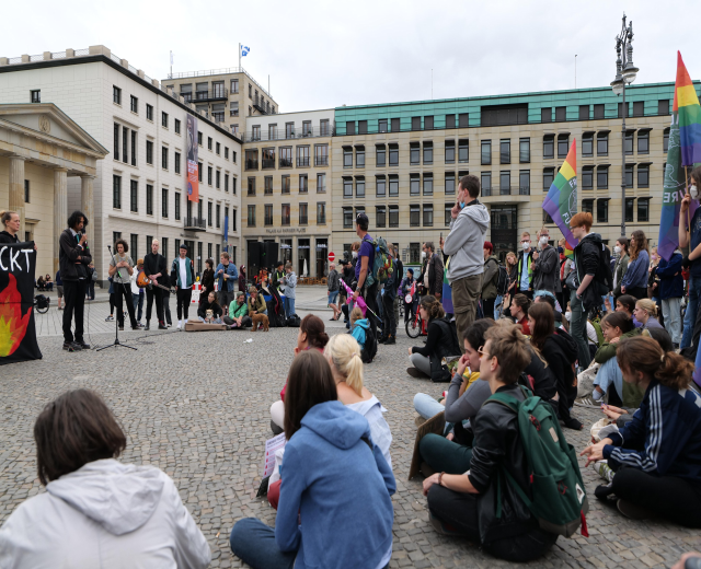 Eine Gruppe von Menschen, die auf dem Boden vor einer Menge sitzen, die Fahnen und Transparente hölt, mit einer Person, die in ein Mikrofon spricht, einer Statue und Gebäuden im Hintergrund bei einer Homo-Protestdemonstration in Berlin.