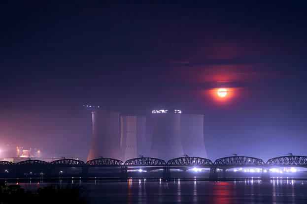 Ein Atomkraftwerk bei Nacht mit einer Brücke über Wasser, beleuchtet von Gebäuden und Mondlicht, umgeben von Bäumen.