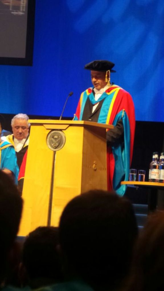 Ein Mann in einer Abschlussrobe und -mütze steht an einem Podium mit einem Mikrofon, mit Menschen darunter, zwei Männern zu seiner Linken und einem Tisch mit Gläsern und Flaschen zu seiner Rechten, vor einem blauen und schwarzen Hintergrund.