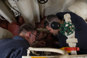 Zwei Männer mit Kopfhörern und Brille arbeiten an einer Maschine in einem Raum voller Rohre, Ventile und anderer Ausrüstung.
