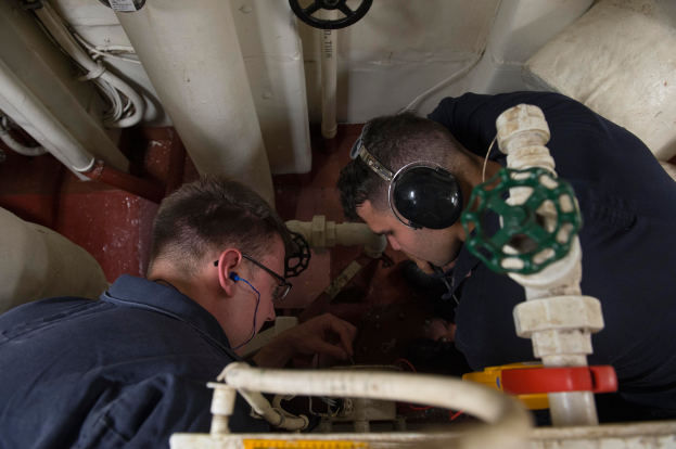 Zwei Männer mit Kopfhörern und Brille arbeiten an einer Maschine in einem Raum voller Rohre, Ventile und anderer Ausrüstung.