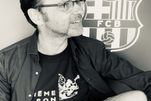 Ein Mann in Brille und schwarzem Hemd lächelt vor einem schwarz-weißen FC Barcelona-Logo an einer Wand.