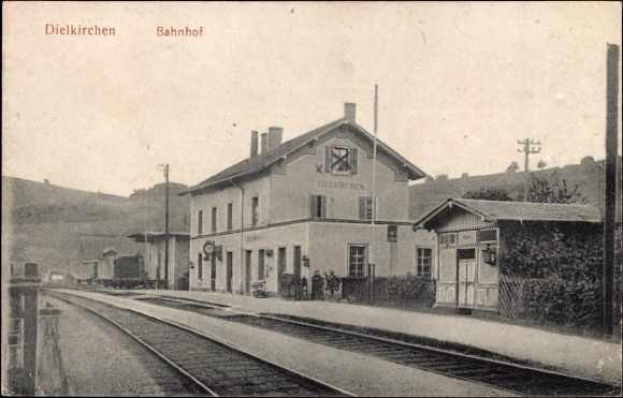 Schwarzes und weißes Foto einer Bahnstation in Dielkirchen, Deutschland, mit einem Zug auf den Schienen, umgeben von Häusern, Pfählen, Bäumen, Pflanzen und Menschen, Bergen im Hintergrund und einem klaren Himmel.