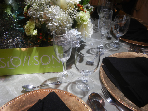 Formaler Esstisch mit goldenen Untersetzern, schwarzen Servietten, Besteck, Gläsern und einem Blumenbouquet, mit einer Texttafel.
