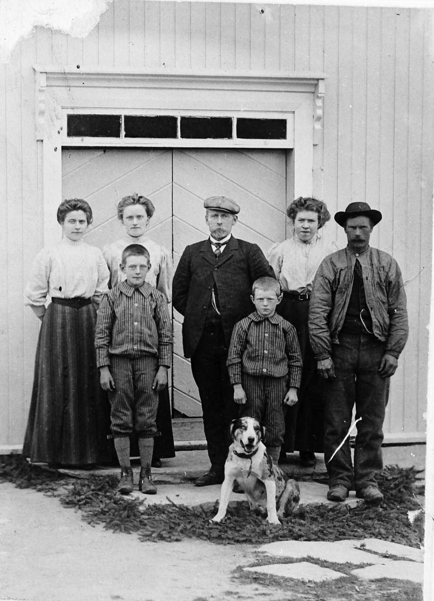 Schwarzes und weißes Foto einer Familie mit einem Hund, die vor einem Haus posieren, bestehend aus einem erwachsenen Mann, einer erwachsenen Frau und mehreren Kindern.