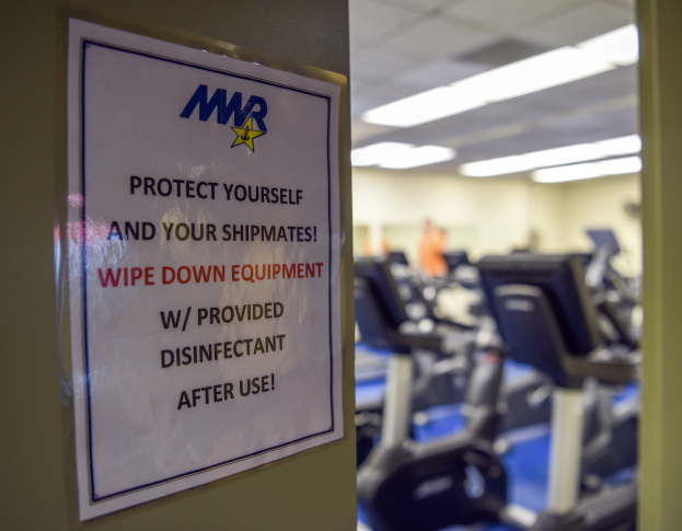 Ein Türschild mit der Aufschrift 'Schützen Sie sich und Ihre Schiffskameraden' mit Laufbändern und Deckenleuchten im Hintergrund, Hintergrund leicht unscharf.