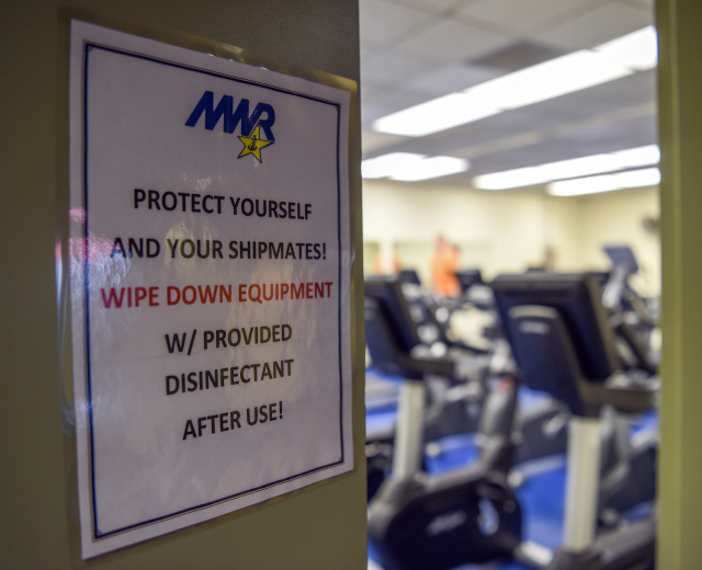 Ein Türschild mit der Aufschrift 'Schützen Sie sich und Ihre Schiffskameraden' mit Laufbändern und Deckenleuchten im Hintergrund, Hintergrund leicht unscharf.