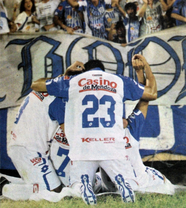 Fußballspieler feiern ein Tor auf dem Feld mit einer jubelnden Menge und einem Banner im Hintergrund.