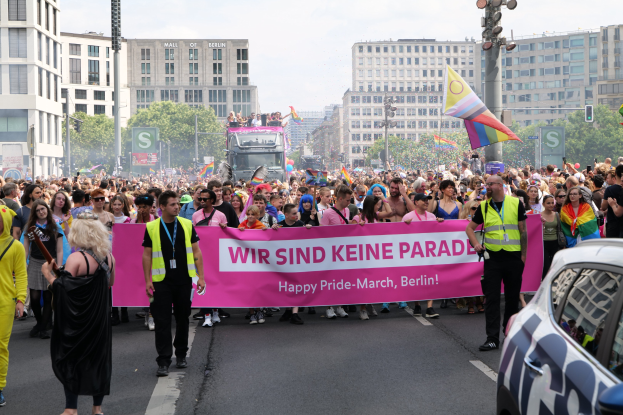 Eine große Gruppe von Menschen marschiert mit einem "Happy Pride March Berlin"-Schild die Straße entlang, mit einem geparkten Auto auf der rechten Seite und Gebäuden, Bäumen und einem klaren Himmel im Hintergrund.
