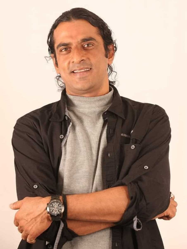 Ein Mann in einem schwarzen und aschfarbenen Kleid mit verschr├Ąnkten Armen, der eine Uhr tr├Ągt, vor einem wei├čem Hintergrund steht und nachdenklich erscheint.