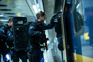 Eine Gruppe von Polizisten steht neben einem Zug auf der rechten Seite des Bildes, wobei einer der Beamten eine Waffe hält und an der Decke befestigte Lampen.