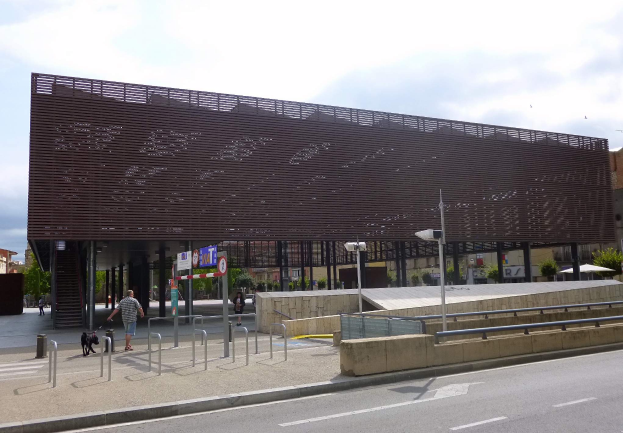 Ein großes Gebäude mit vielen Fenstern, das als neuer Eingang zur Stadt dient, umgeben von Straßenlaternen, Fahrzeugen, Fußgängern, Sonnenschirmen, Bäumen und einem bewölkten Himmel.