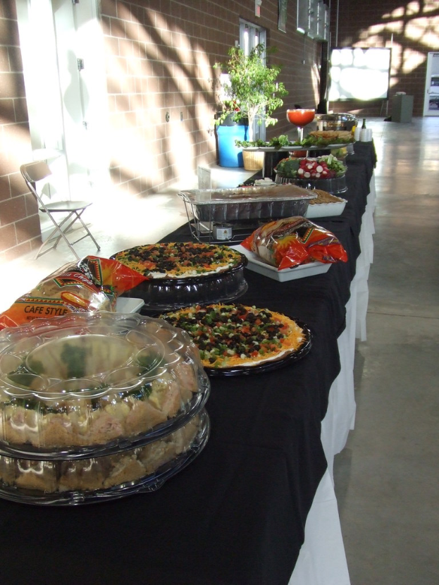 Ein Tisch mit einer schwarzen und weißen Decke darauf, auf dem ein Teller mit Pizza, ein Tablett mit Plastiktüten und Gefäße mit Essen stehen; daneben ein Stuhl, mit einem braunen Gebäude, einem Blumentopf und einem blauen Müllcontainer im Hintergrund.