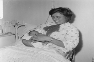 Eine Frau in einem Krankenhausbett hält ein Neugeborenes im Arm, umgeben von einem Kissen und einer Decke, mit einem Tisch links und Rohren im Hintergrund.