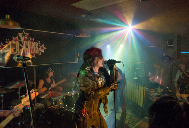 Eine Frau in einer schwarzen Jacke singt in ein Mikrofon auf der Bühne, mit einem Mann in Sonnenbrille und einem Stirnband, der ein Instrument spielt, und einem Plakat mit "LA Guns" im Hintergrund.