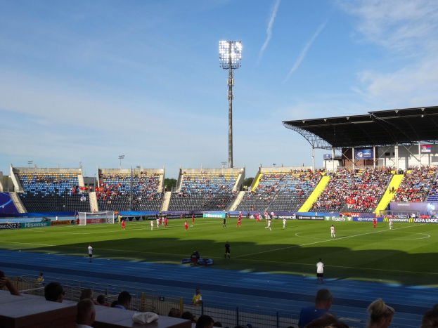 Ein Fußballspiel in einem großen Stadion mit Spielern auf dem Feld und Zuschauern auf den Rängen unter Flutlicht, mit dem Himmel im Hintergrund.