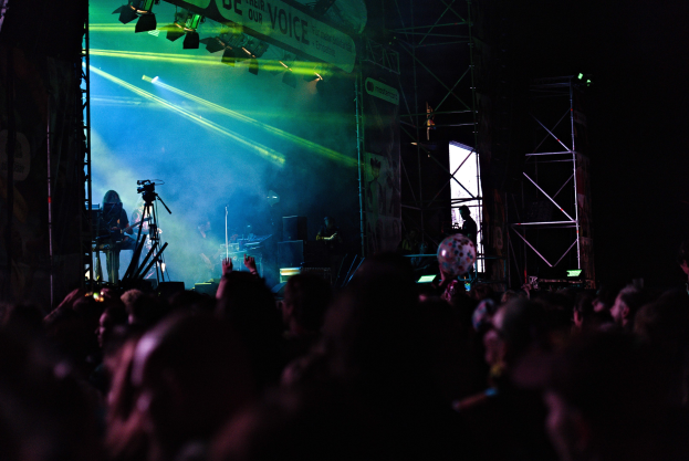 Ein volles Konzert mit Musikern auf der Bühne unter Scheinwerfern, Metallrahmen und einem Banner im Hintergrund.