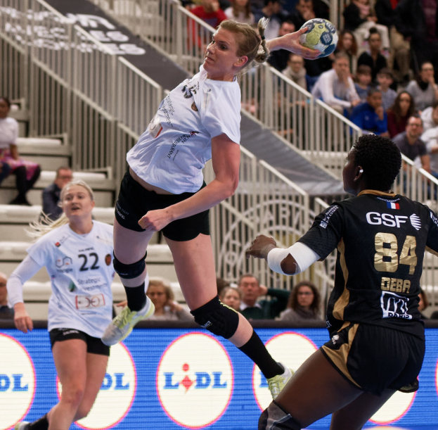 Frauen spielen Handball auf einem Court mit einem Ball in der Luft, Zuschauer im Hintergrund und einem "Handball-Weltmeisterschaft 2019 - Tag 2"-Schild.