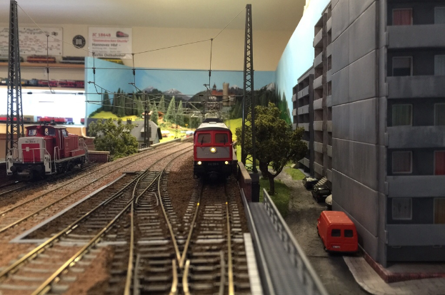 Ein Spielzeughahn und eine Lokomotive auf Schienen mit Bäumen, Pfählen, Kabeln, Gebäuden, Fahrzeugen, Geländern, Himmel, Bergen, Grasland und Pfosten im Hintergrund.