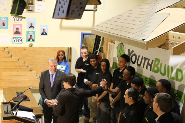 Eine Gruppe von Menschen steht um ein Rednerpult vor einem Publikum, einige schütteln Hände, ein Tisch mit Mikrofonen und Papieren links, Plakate an der Wand im Hintergrund und Gegenstände auf dem Dach und der rechten Seite.