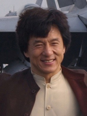 Jackie Chan steht lachend vor einem Flugzeug mit einem klaren blauen Himmel im Hintergrund, trägt ein weißes Hemd, eine schwarze Jacke und hat ordentlich frisiertes Haar.