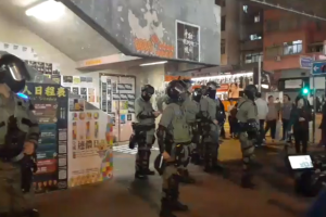 Eine Gruppe von Polizisten in Uniform steht vor einer Menge von Demonstranten mit Helmen und Waffen, mit Plakaten an den Wänden, Lichtern an der Decke und Gebäuden mit Fenstern im Hintergrund, in Hong Kong.