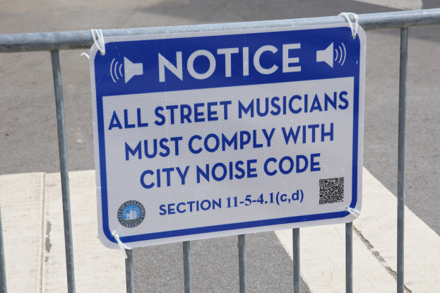 Anschlagtafel mit einem Schild, auf dem "Anschlag: Alle Straßenmusiker müssen sich an die Stadtlärmvorschrift halten" steht, an einem Zaun befestigt, mit einer Straße im Hintergrund.